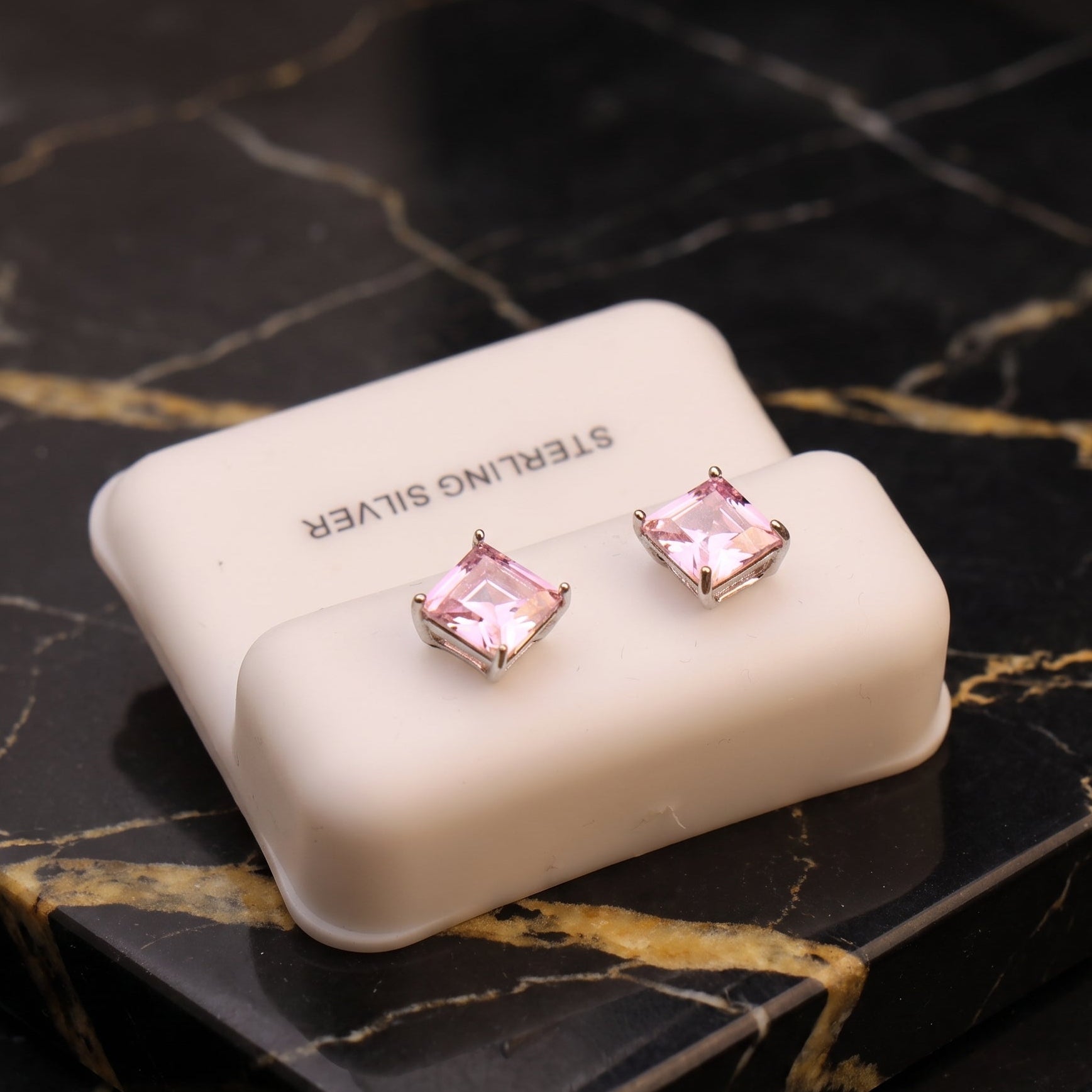 A pair of silver stud earrings with square pink gemstones, displayed on a white 'STERLING SILVER' box set on a black marble surface with gold veining.