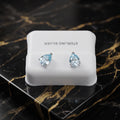 A pair of silver stud earrings with pear-cut light blue gemstones, displayed on a white jewelry box labeled 'STERLING SILVER' on a black and gold marble surface.