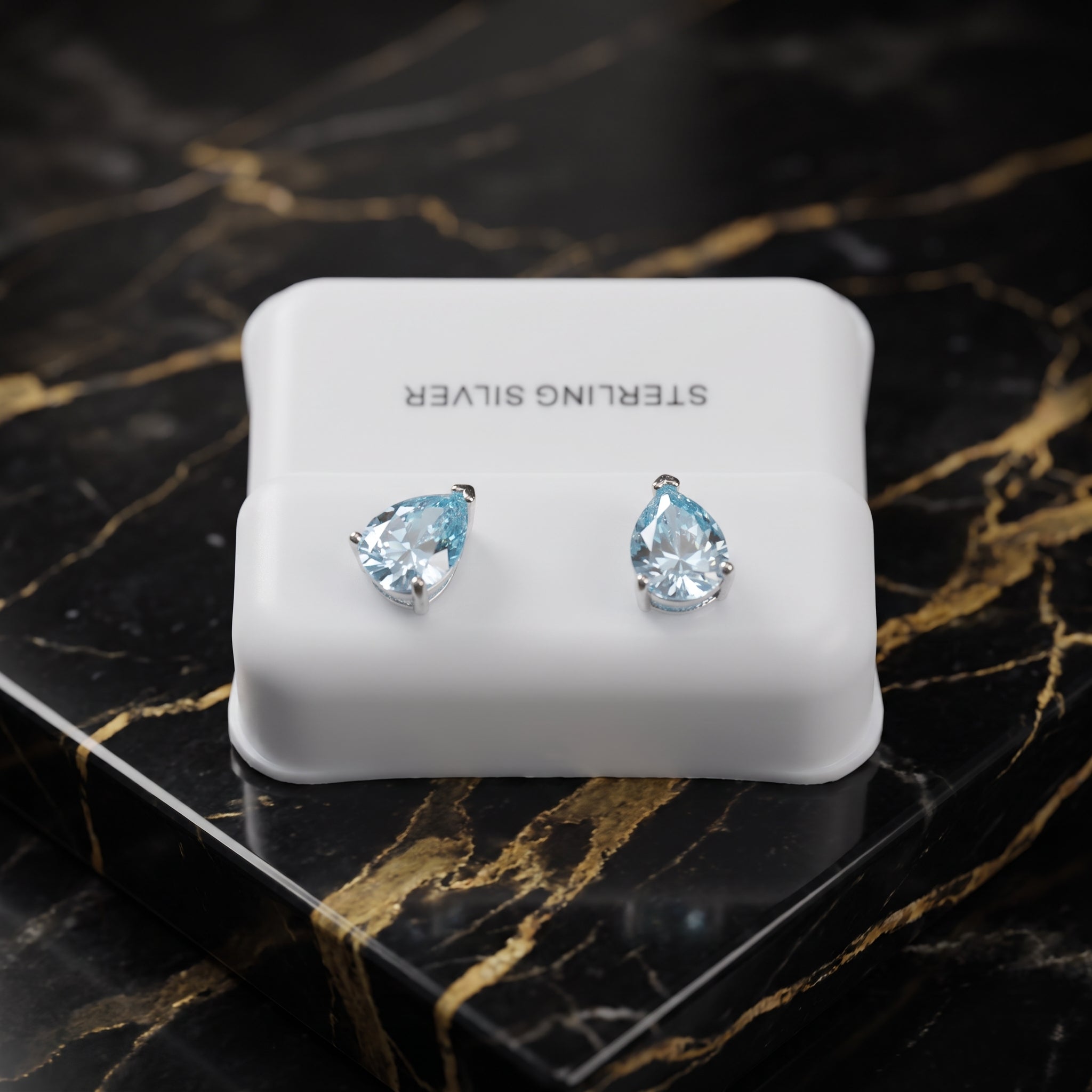 A pair of silver stud earrings with pear-cut light blue gemstones, displayed on a white jewelry box labeled 'STERLING SILVER' on a black and gold marble surface.