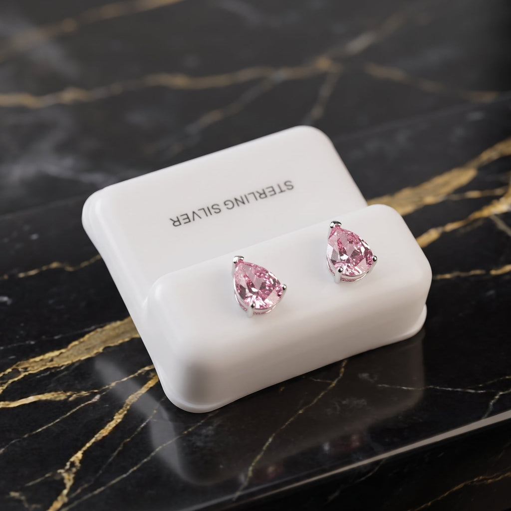 A pair of silver stud earrings with teardrop-shaped pink gemstones, displayed on a white jewelry stand labeled 'STERLING SILVER' on a black marble surface with gold veining.