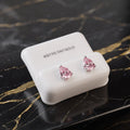 A pair of silver stud earrings with teardrop-shaped pink gemstones, displayed on a white jewelry stand labeled 'STERLING SILVER' on a black marble surface with gold veining.