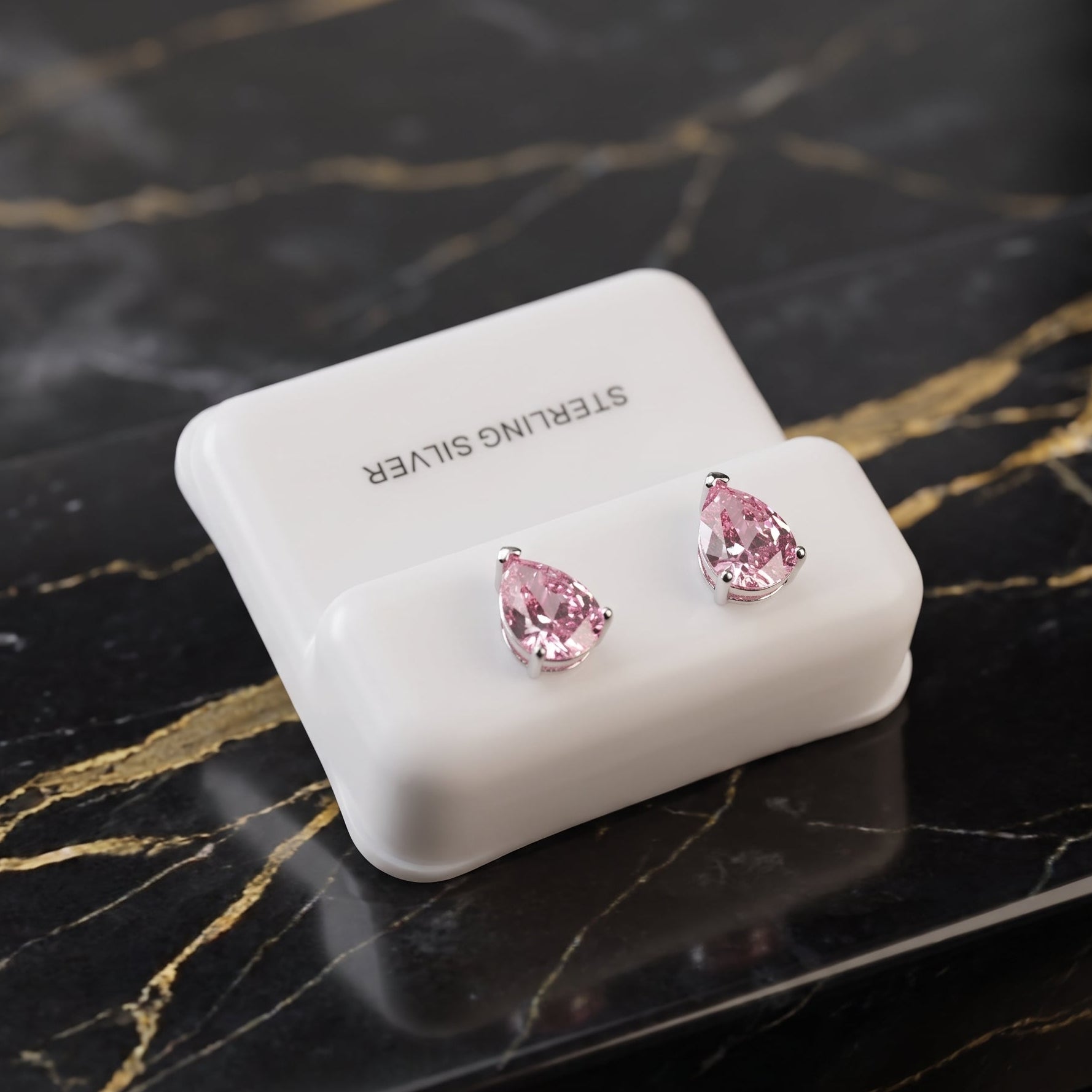 A pair of silver stud earrings with teardrop-shaped pink gemstones, displayed on a white jewelry stand labeled 'STERLING SILVER' on a black marble surface with gold veining.