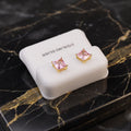 A pair of gold stud earrings with pink square-cut stones, displayed on a white jewelry stand labeled 'STERLING SILVER' on a black marble surface with gold veining.