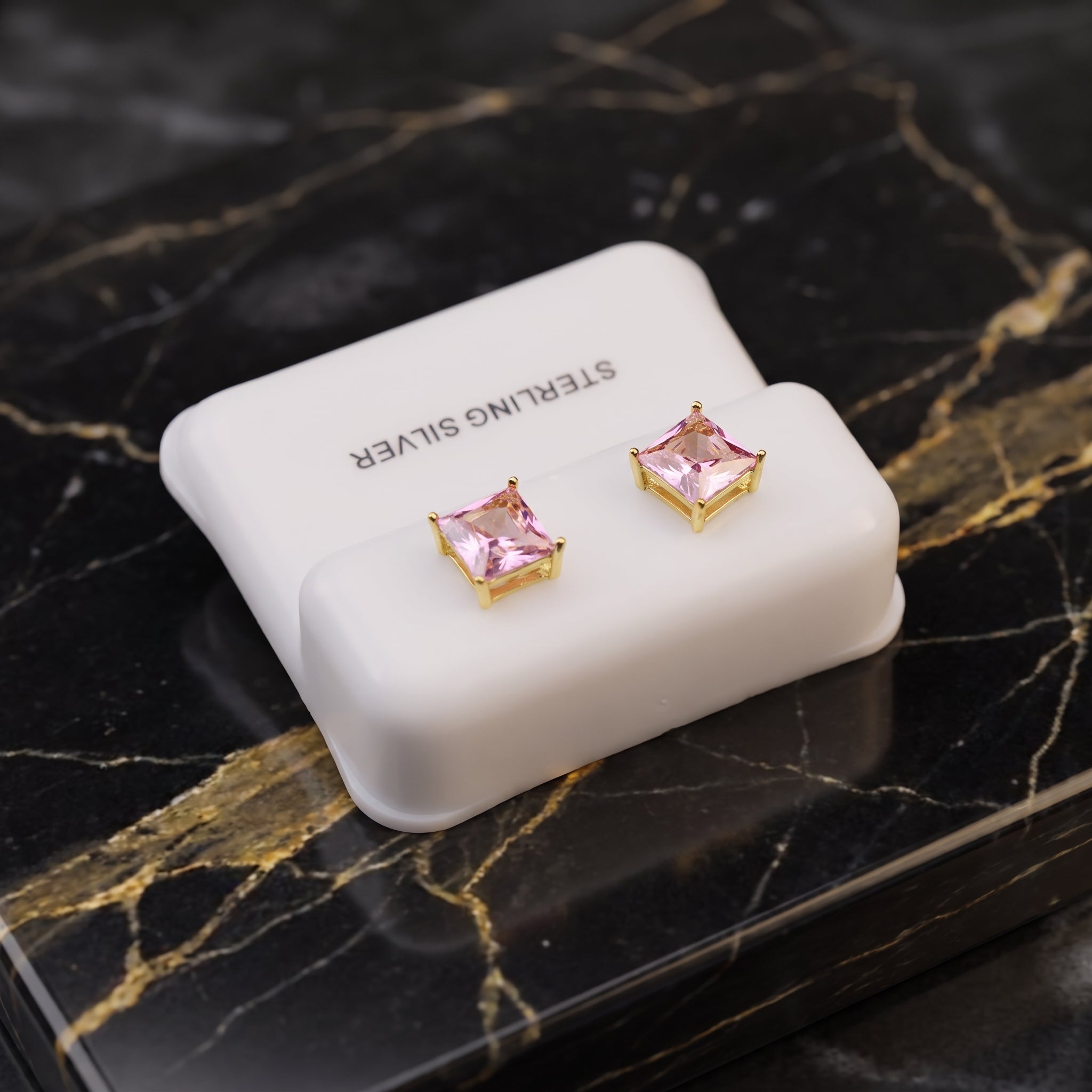 A pair of gold stud earrings with pink square-cut stones, displayed on a white jewelry stand labeled 'STERLING SILVER' on a black marble surface with gold veining.