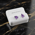 A pair of silver stud earrings with teardrop-shaped purple gemstones, displayed on a white jewelry box set against a black marble background with gold veining.