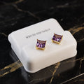 A pair of square stud earrings with purple gemstones in gold settings, displayed on a white jewelry box labeled 'STERLING SILVER' on a black marble surface.