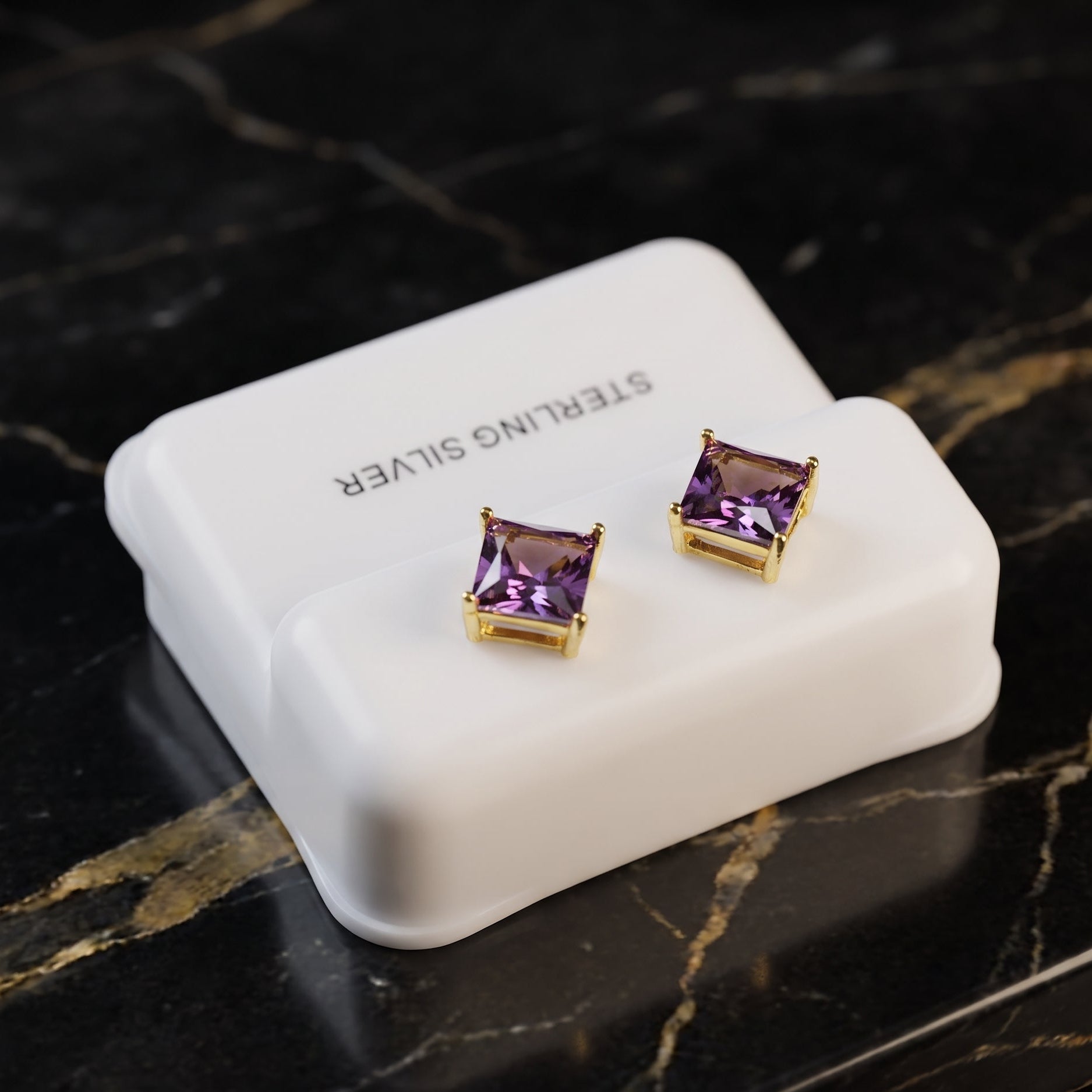 A pair of square stud earrings with purple gemstones in gold settings, displayed on a white jewelry box labeled 'STERLING SILVER' on a black marble surface.