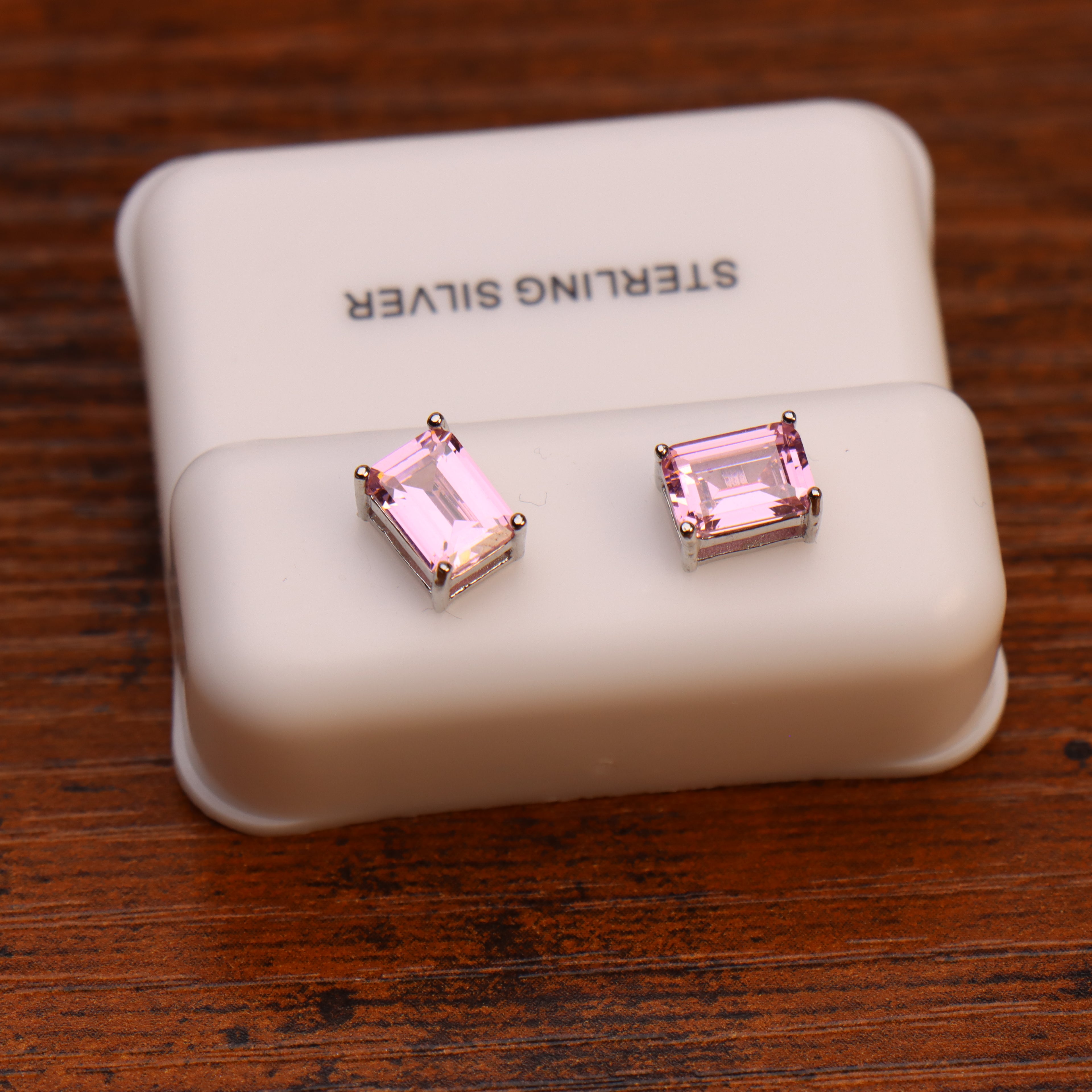 A pair of emerald cut pink sapphire cubic zirconia earrings (7x5mm) set in 925 sterling silver, displayed on a white jewelry holder labeled STERLING SILVER atop a wooden surface.