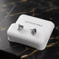 A pair of heart-shaped stud earrings with iridescent faceted stones in silver settings, displayed on a white rectangular jewelry box that reads 'STERLING SILVER,' placed on a black marble surface with gold veining.