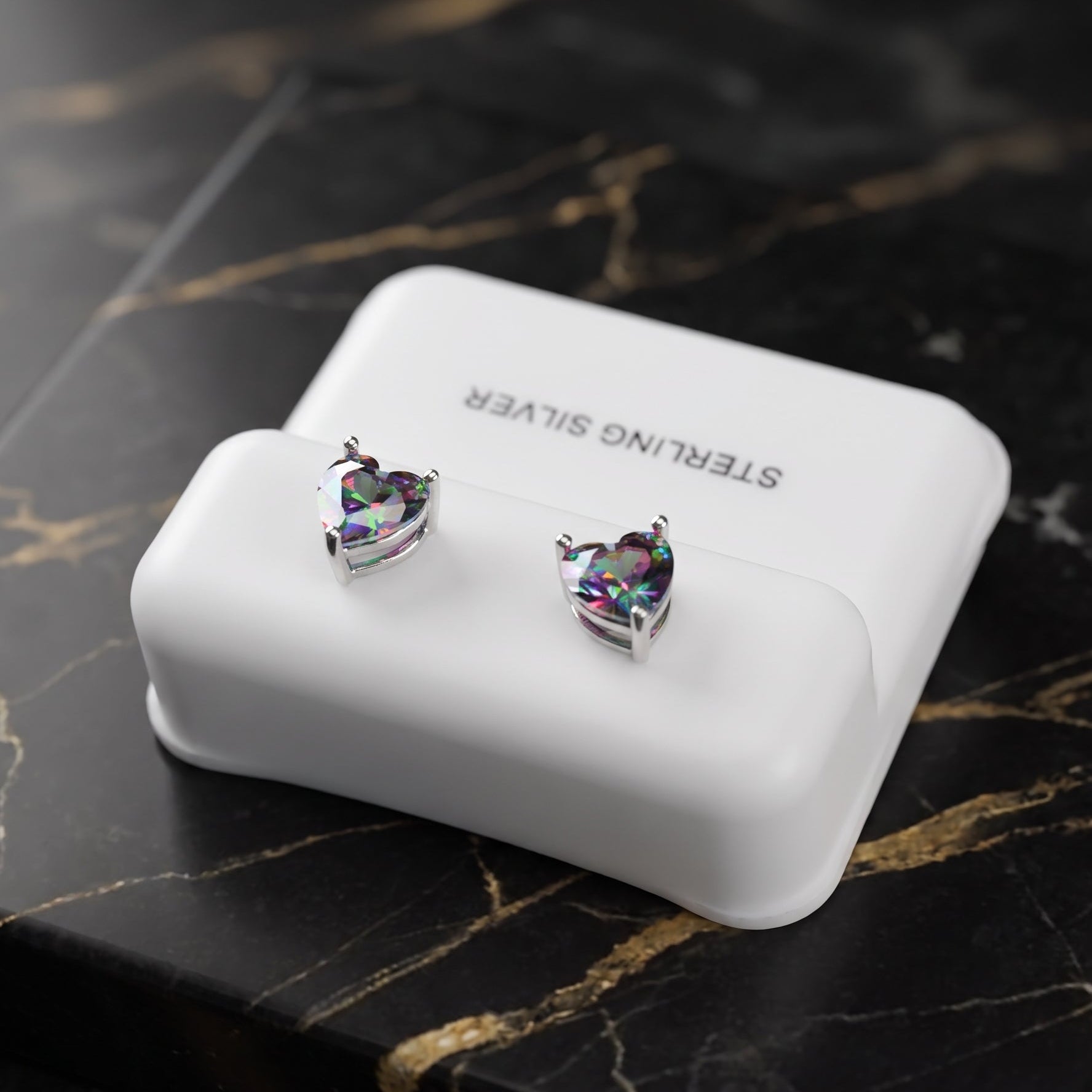 A pair of heart-shaped stud earrings with iridescent faceted stones in silver settings, displayed on a white rectangular jewelry box that reads 'STERLING SILVER,' placed on a black marble surface with gold veining.