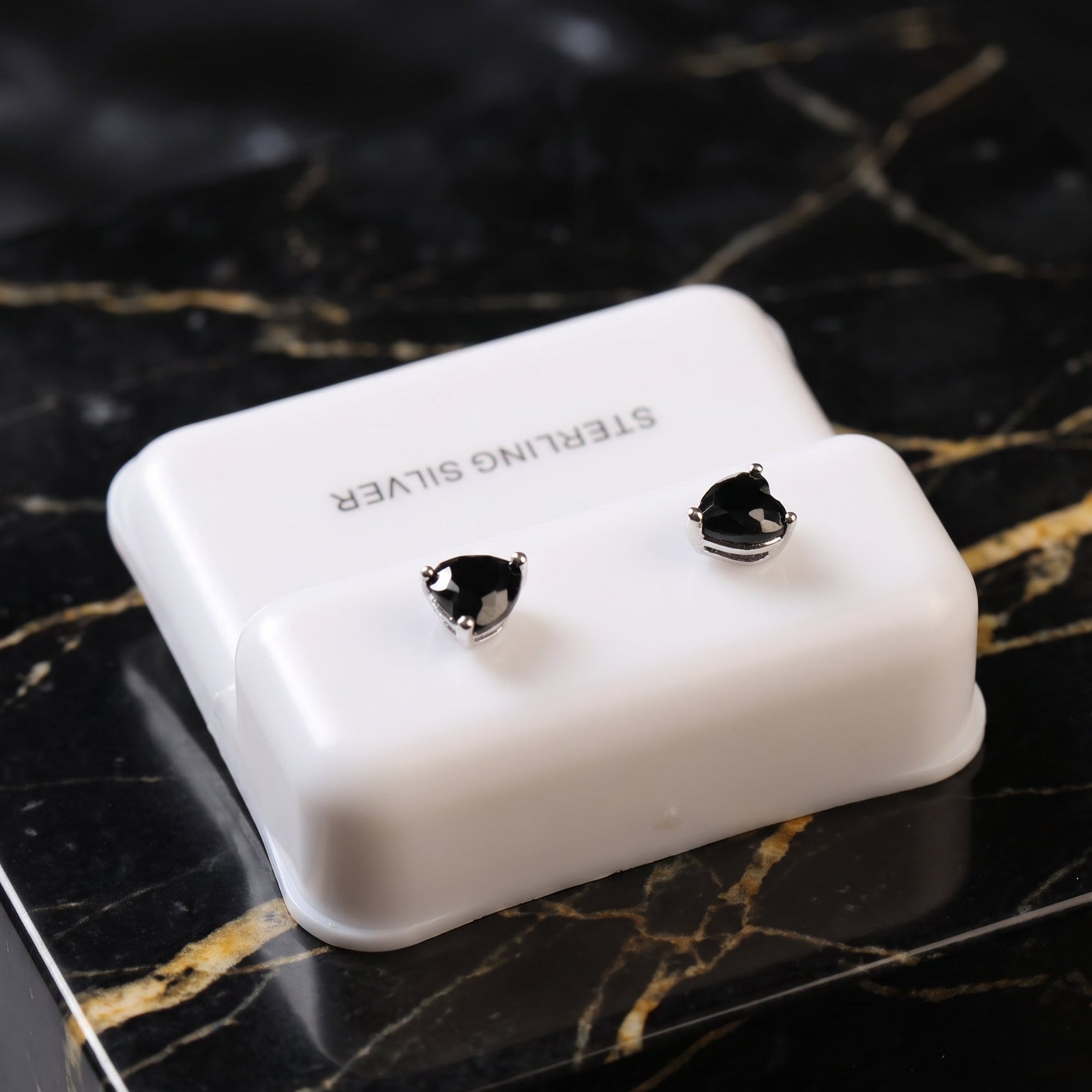 A pair of sterling silver stud earrings with black heart-shaped stones, displayed on a white plastic jewelry stand set on a black marble surface.