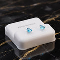 A pair of small blue teardrop-shaped stud earrings displayed on a white rectangular jewelry box, set on a black marble surface with gold veining.