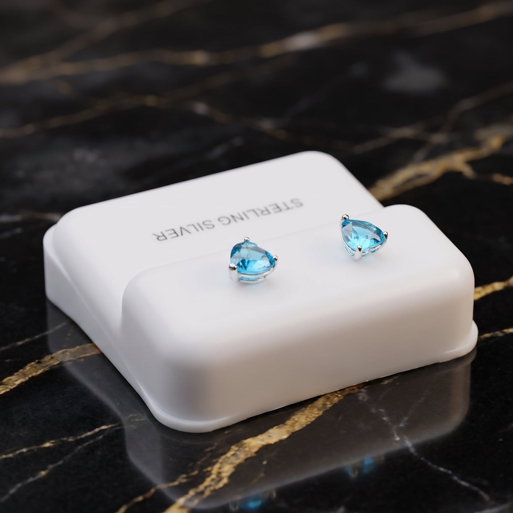 A pair of small blue teardrop-shaped stud earrings displayed on a white rectangular jewelry box, set on a black marble surface with gold veining.