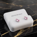 A pair of silver stud earrings with pink heart-shaped gemstones, displayed on a white rectangular jewelry box labeled 'STERLING SILVER' on a black marble surface with gold veining.