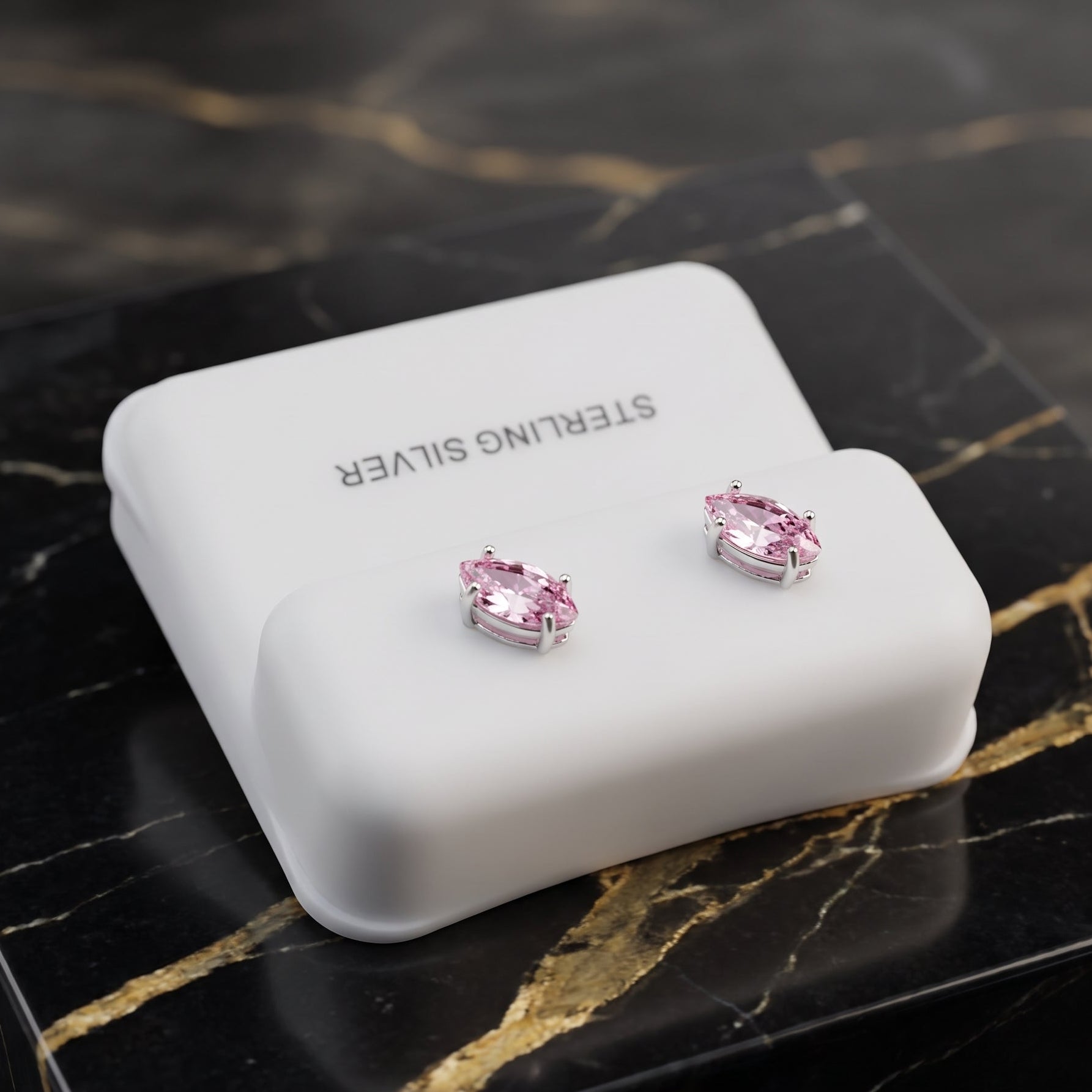A pair of silver stud earrings with pink heart-shaped gemstones, displayed on a white rectangular jewelry box labeled 'STERLING SILVER' on a black marble surface with gold veining.