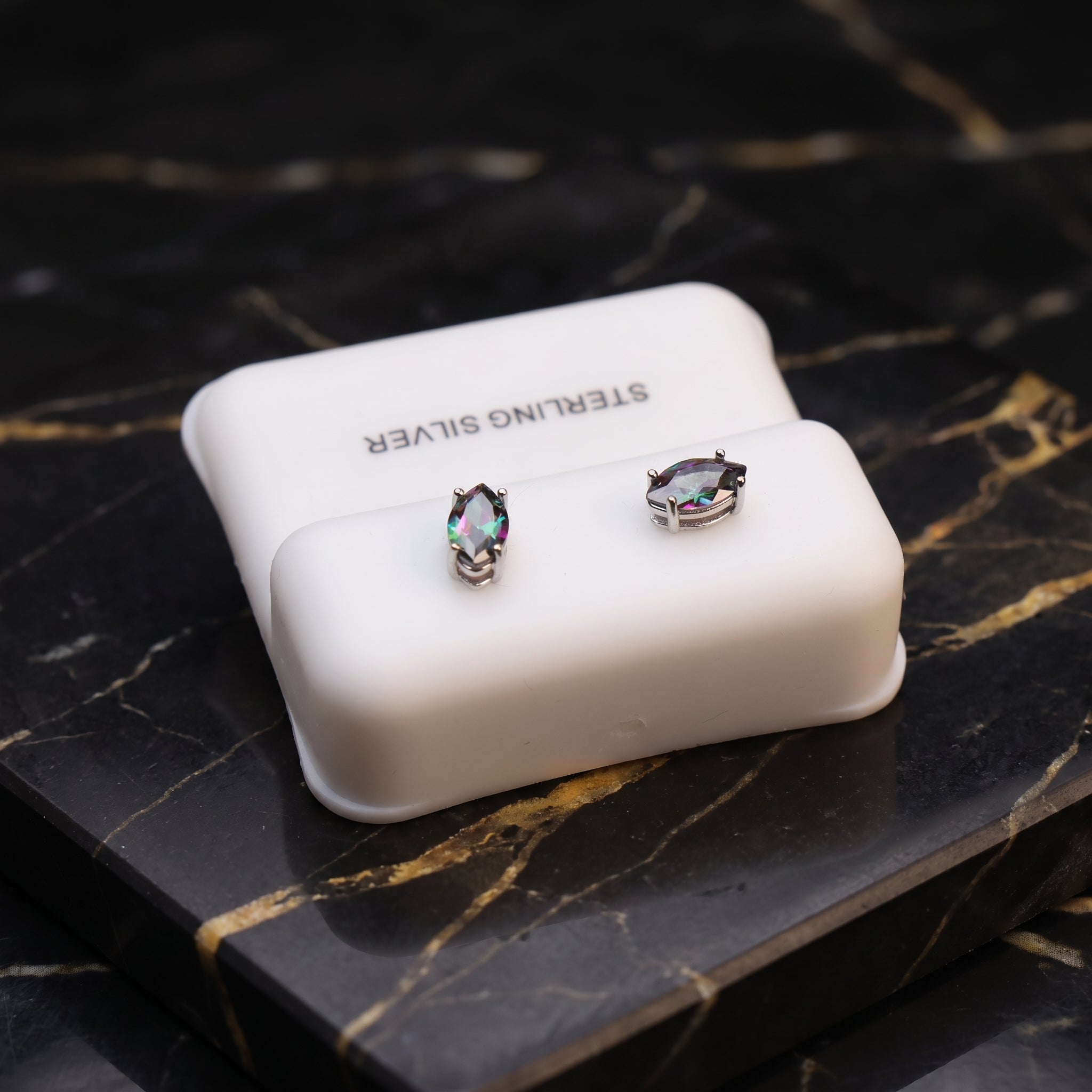 A pair of oval-shaped sterling silver stud earrings with iridescent, rainbow-like stones, displayed on a white rectangular jewelry box labeled 'STERLING SILVER' on a black marble surface with gold veining.