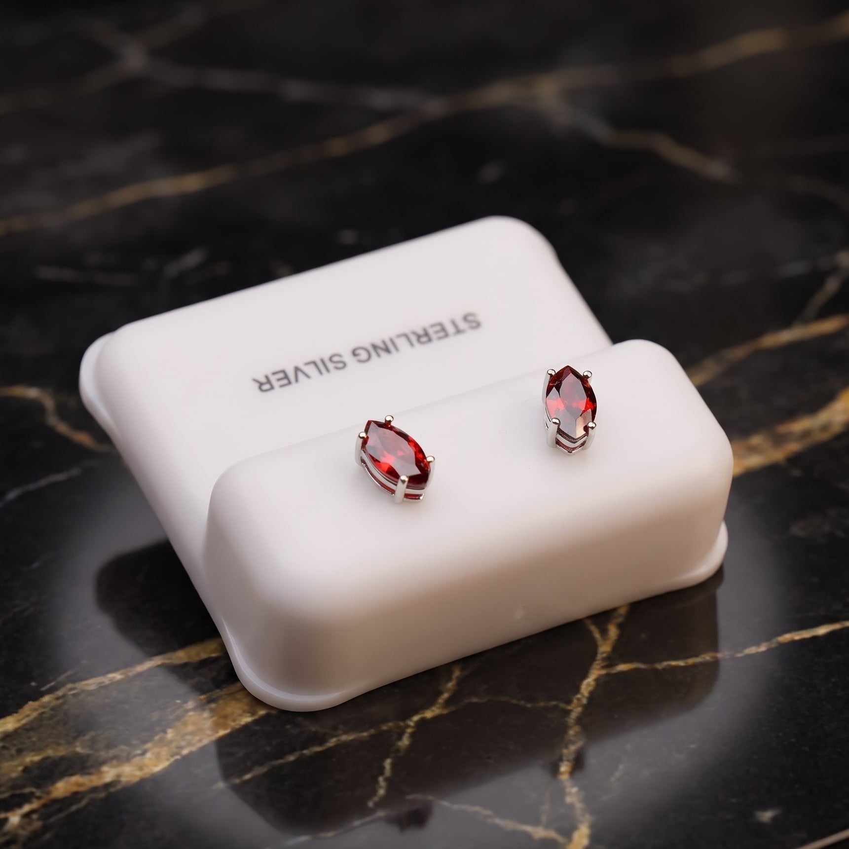 A pair of silver stud earrings with marquise-cut red gemstones displayed on a white jewelry stand, set on a black marble surface.