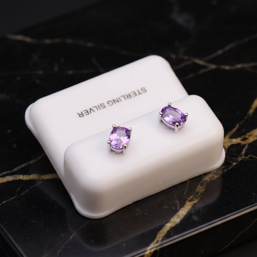 A pair of oval-cut purple gemstone stud earrings in silver settings, displayed on a white cushion atop a black marble surface. The cushion is printed with the text 'STERLING SILVER'.