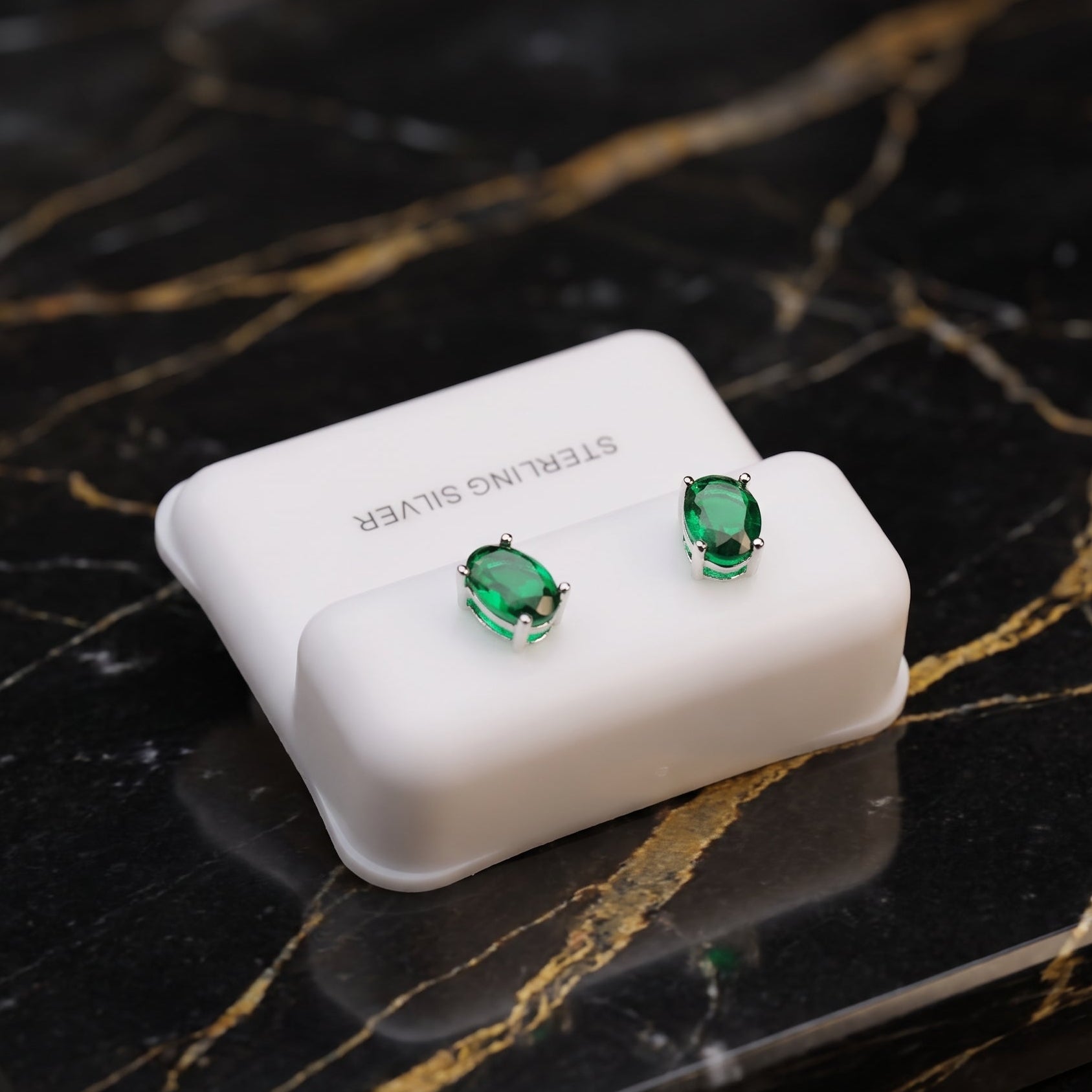 A pair of oval green gemstone stud earrings displayed on a white jewelry box labeled 'STERLING Silver,' placed on a black marble surface with gold veining.