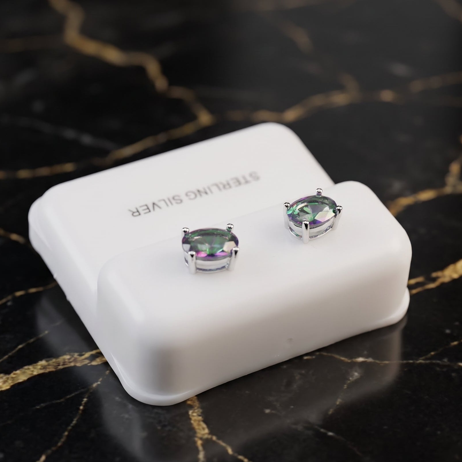 A pair of silver stud earrings with oval iridescent green-purple stones, displayed on a white jewelry stand labeled 'STERLING SILVER' on a black marble surface with gold veining.