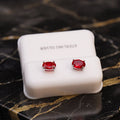 A pair of silver stud earrings with oval red gemstones, displayed on a white jewelry box labeled 'STERLING SILVER' on a black marble surface with gold veining.