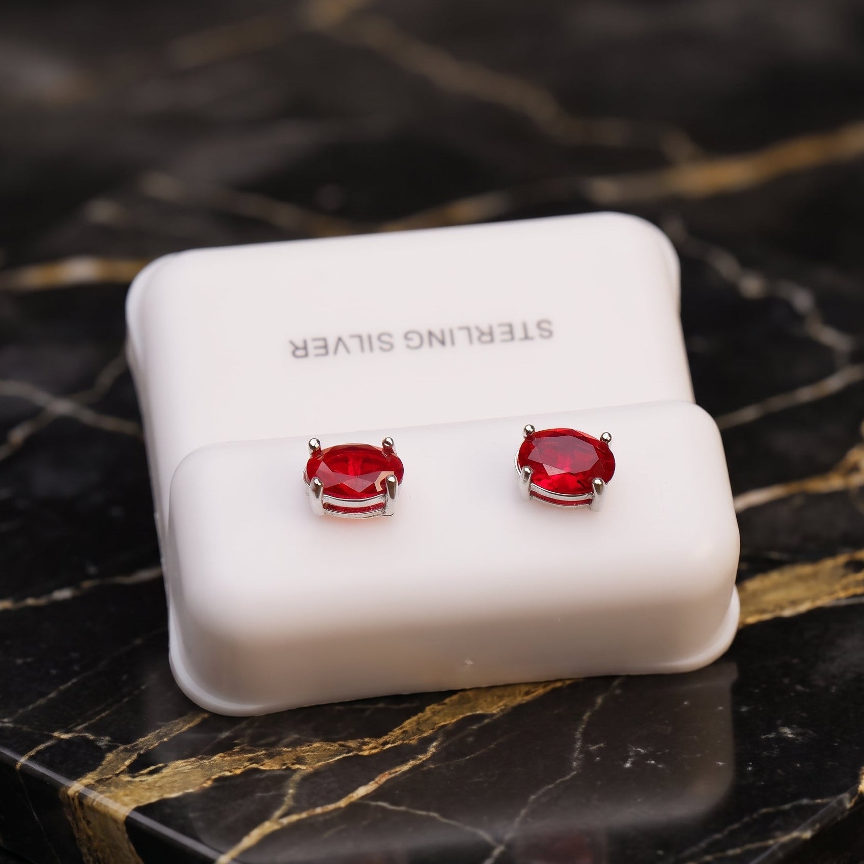 A pair of silver stud earrings with oval red gemstones, displayed on a white jewelry box labeled 'STERLING SILVER' on a black marble surface with gold veining.