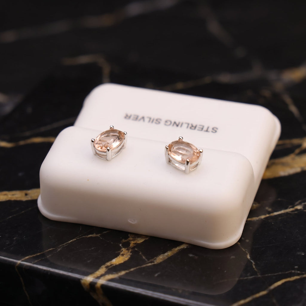 A pair of oval-cut silver stud earrings with clear stones, displayed on a white jewelry box labeled 'STERLING SILVER' on a black marble surface. morganite