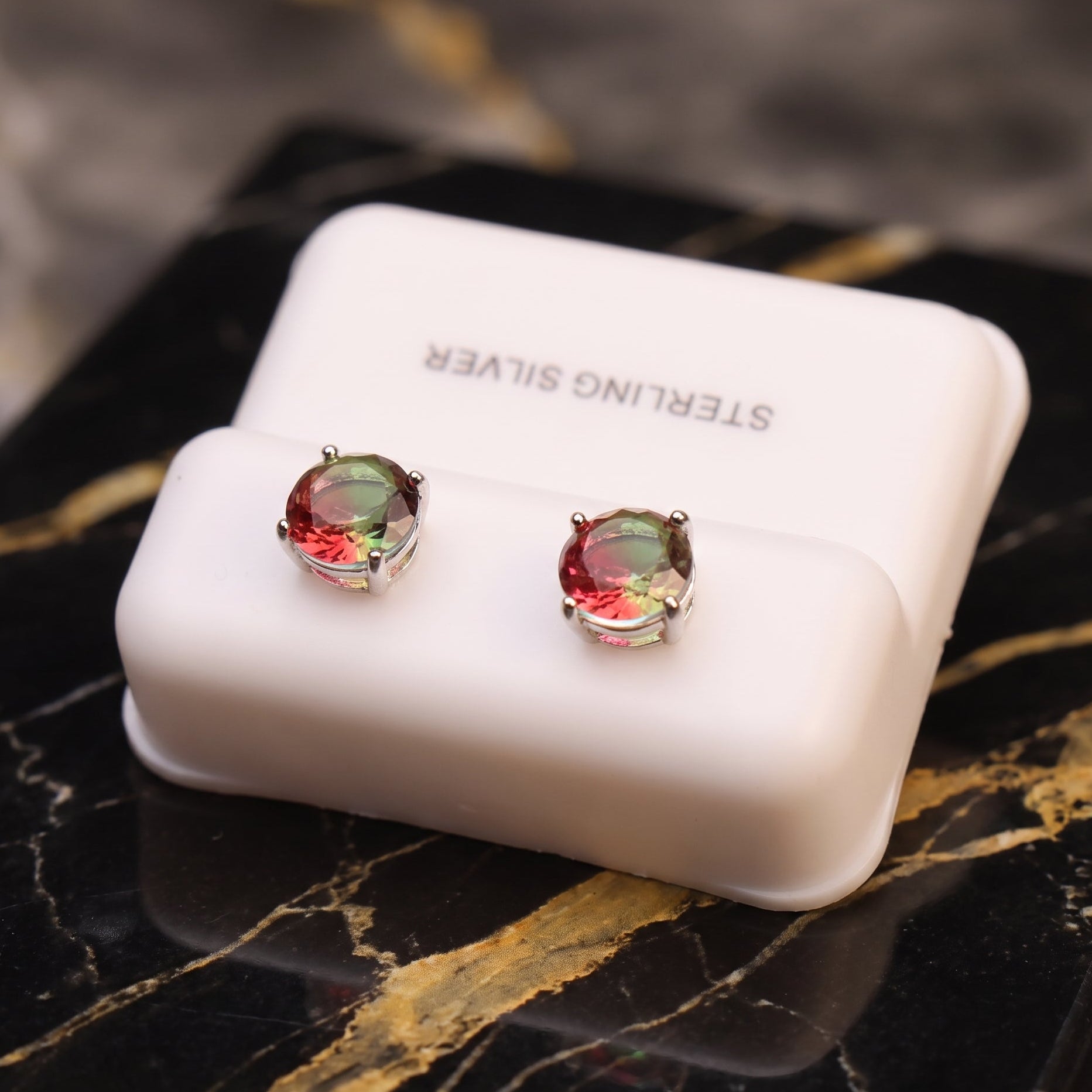 A pair of watermelon round-cut gemstone stud earrings displayed on a white jewelry box labeled 'STERLING SILVER,' set on a black marble surface with gold veining.