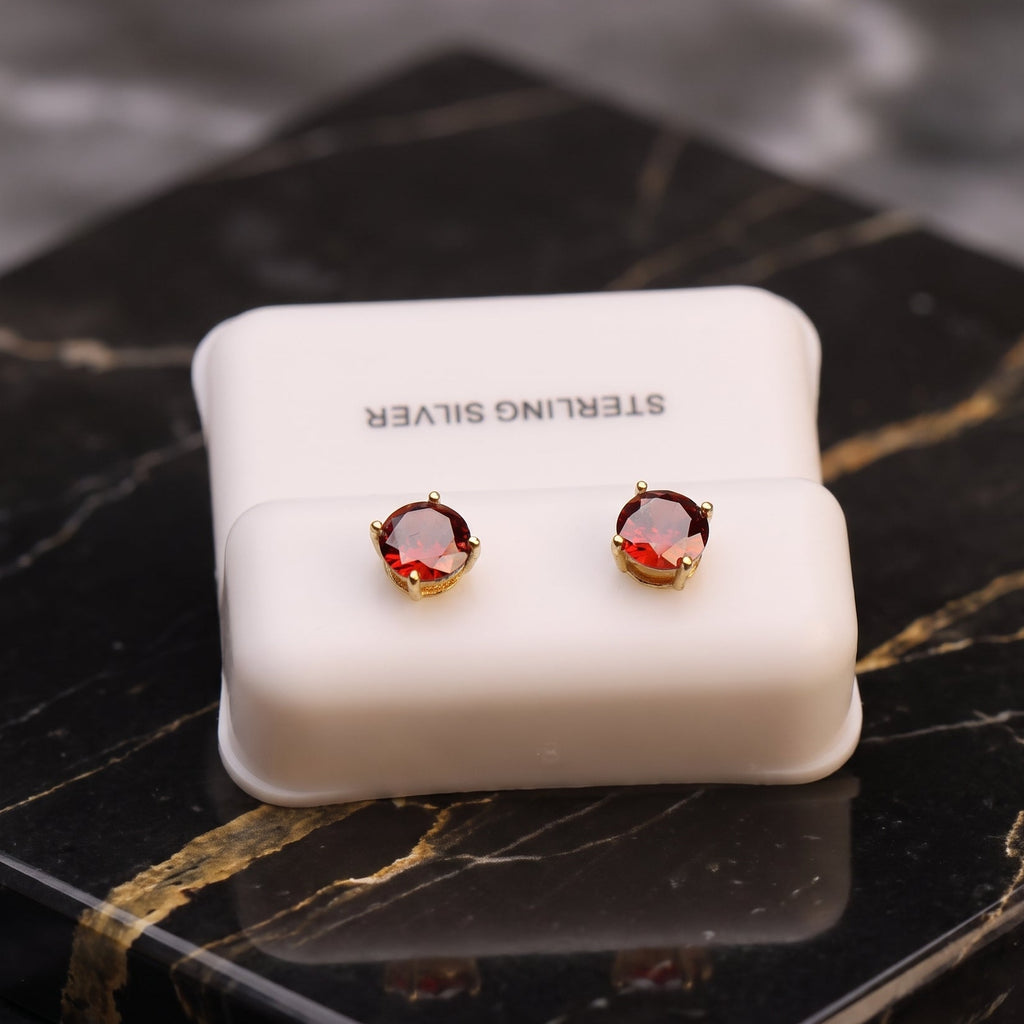 A pair of round stud earrings with deep red gemstones in gold settings, displayed on a white jewelry box labeled 'STERLING SILVER' on a black marble surface.