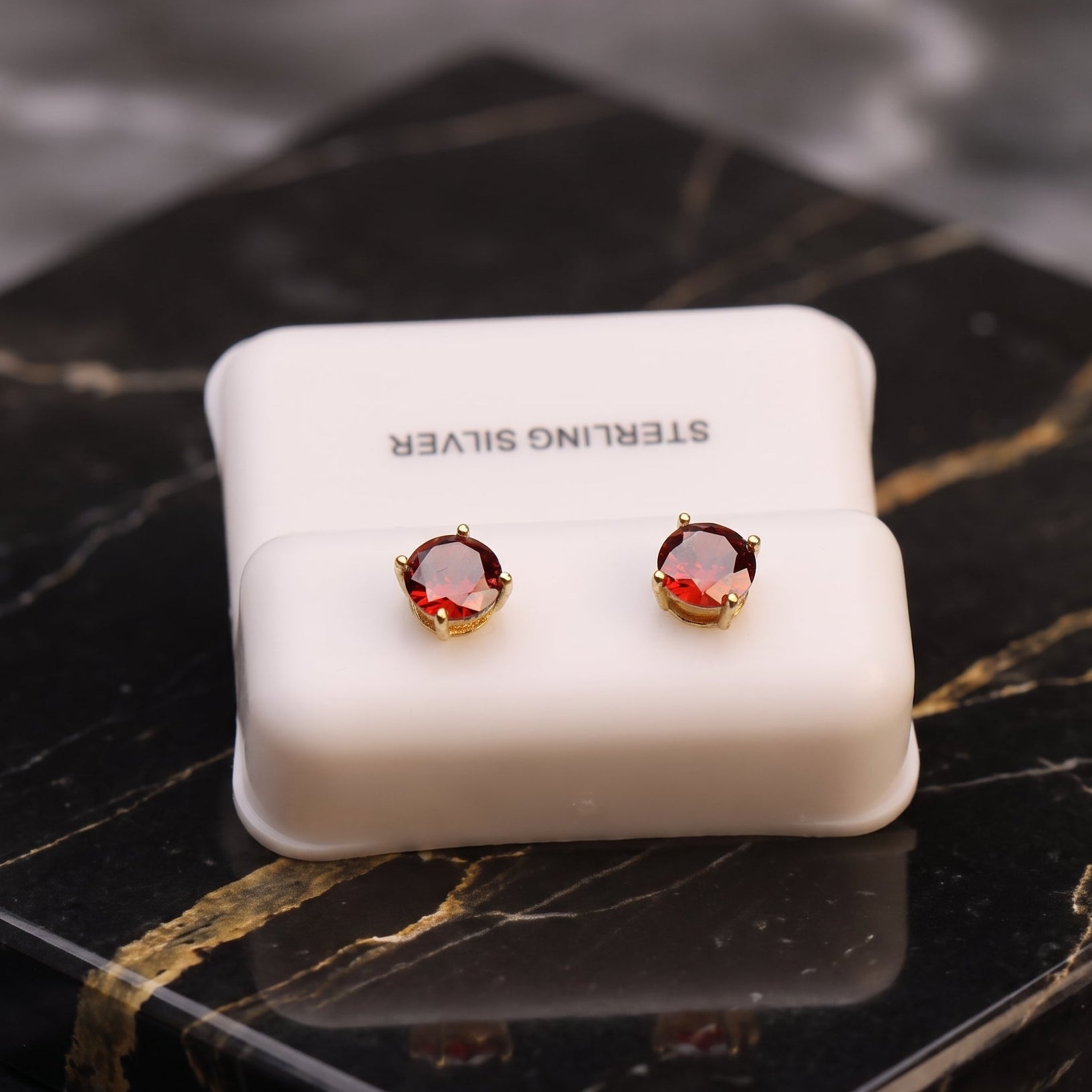 A pair of round stud earrings with deep red gemstones in gold settings, displayed on a white jewelry box labeled 'STERLING SILVER' on a black marble surface.