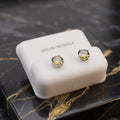 A pair of stud earrings displayed on a white rectangular jewelry box, placed on a black marble surface with gold veining. The earrings have clear faceted round stones in gold-toned settings with silver-toned prongs.