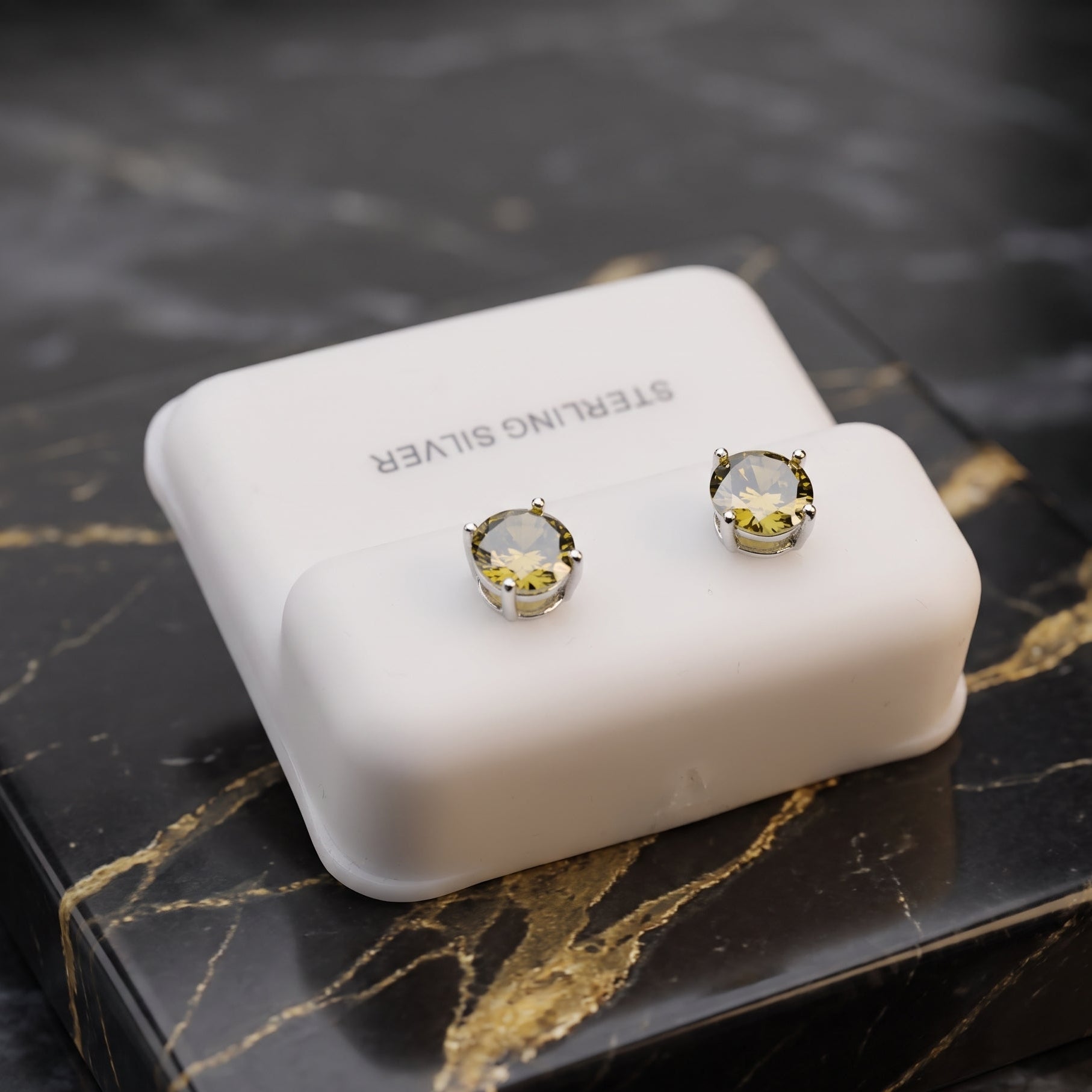 A pair of stud earrings displayed on a white rectangular jewelry box, placed on a black marble surface with gold veining. The earrings have clear faceted round stones in gold-toned settings with silver-toned prongs.