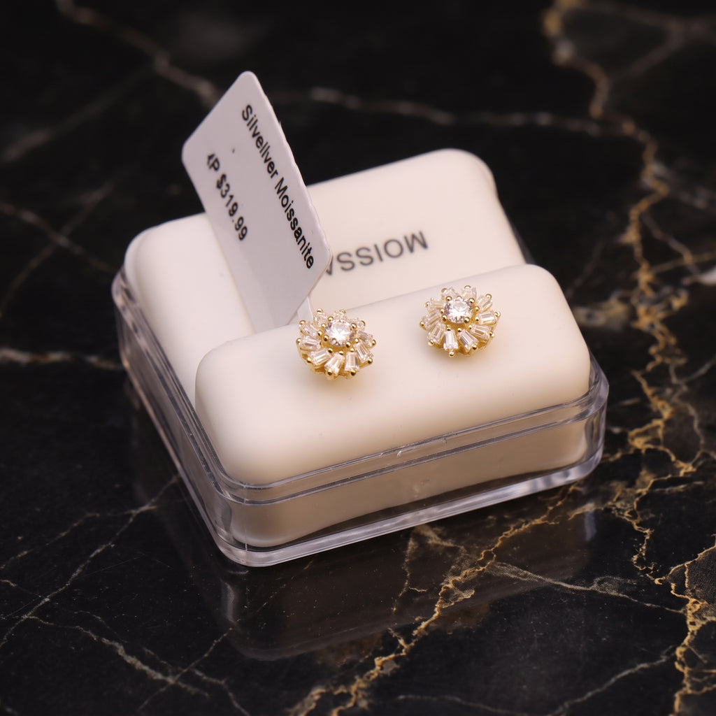 A pair of gold flower-shaped stud earrings with clear gemstones, displayed on a white cushion inside a clear jewelry box on a black marble surface.
