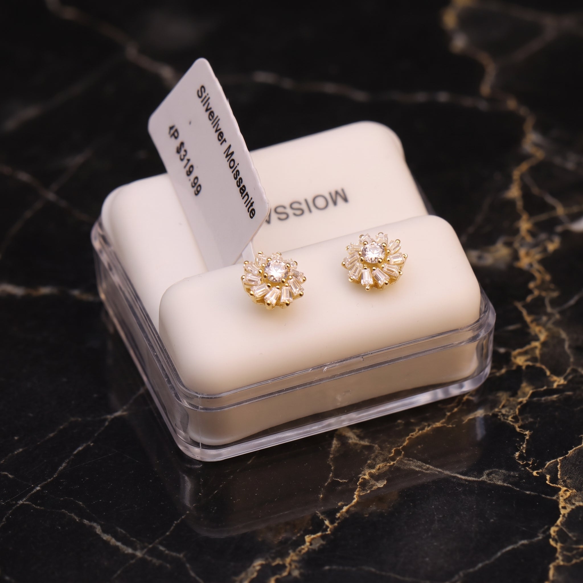 A pair of gold flower-shaped stud earrings with clear gemstones, displayed on a white cushion inside a clear jewelry box on a black marble surface.