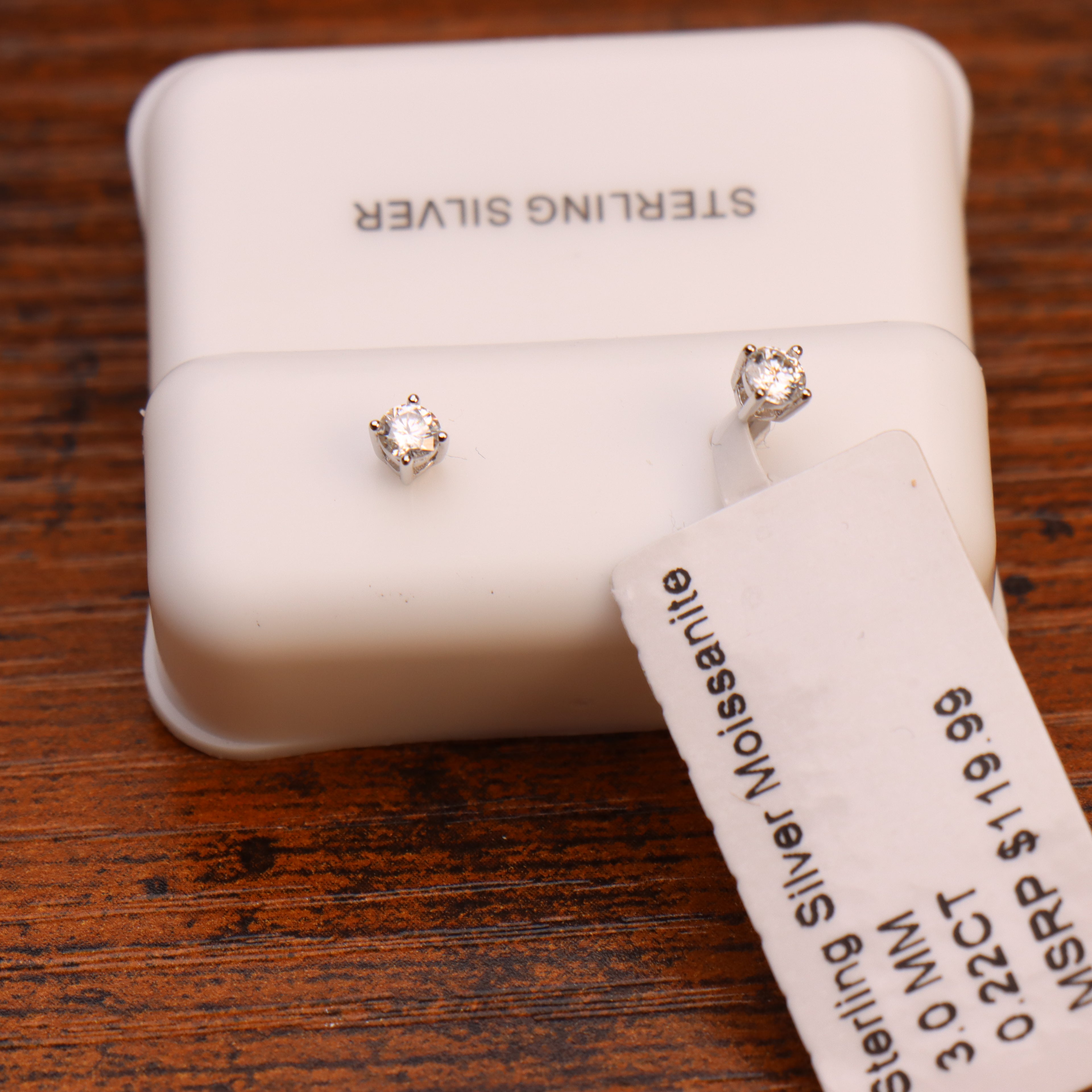 A pair of small silver stud earrings with clear gemstones displayed on a white jewelry box, with a price tag attached and the words 'STERLING SILVER' visible on the box and tag.