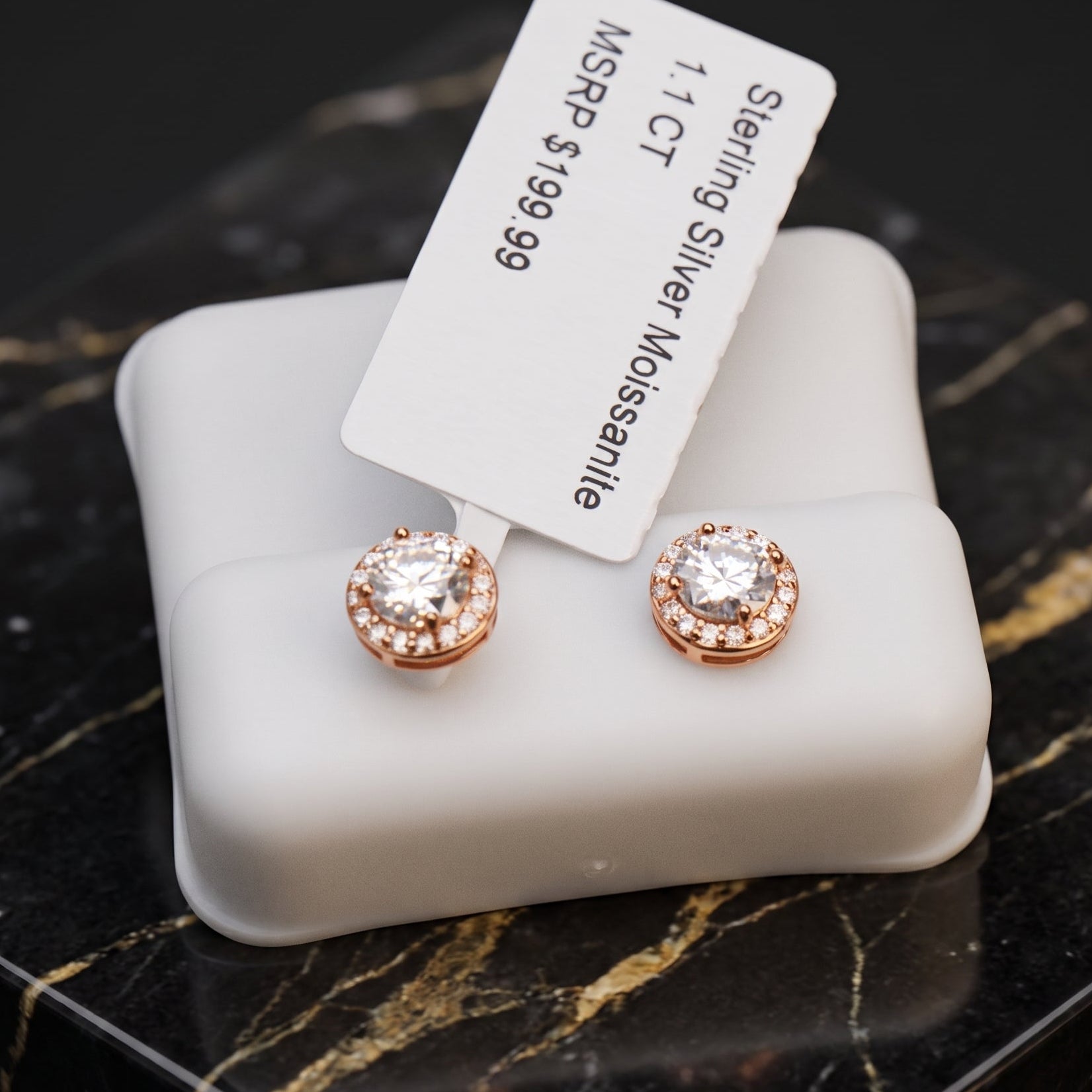A pair of stud earrings displayed on a white jewelry box, with a white tag attached. The earrings have rose gold-toned settings with small clear stones around a larger clear center stone.