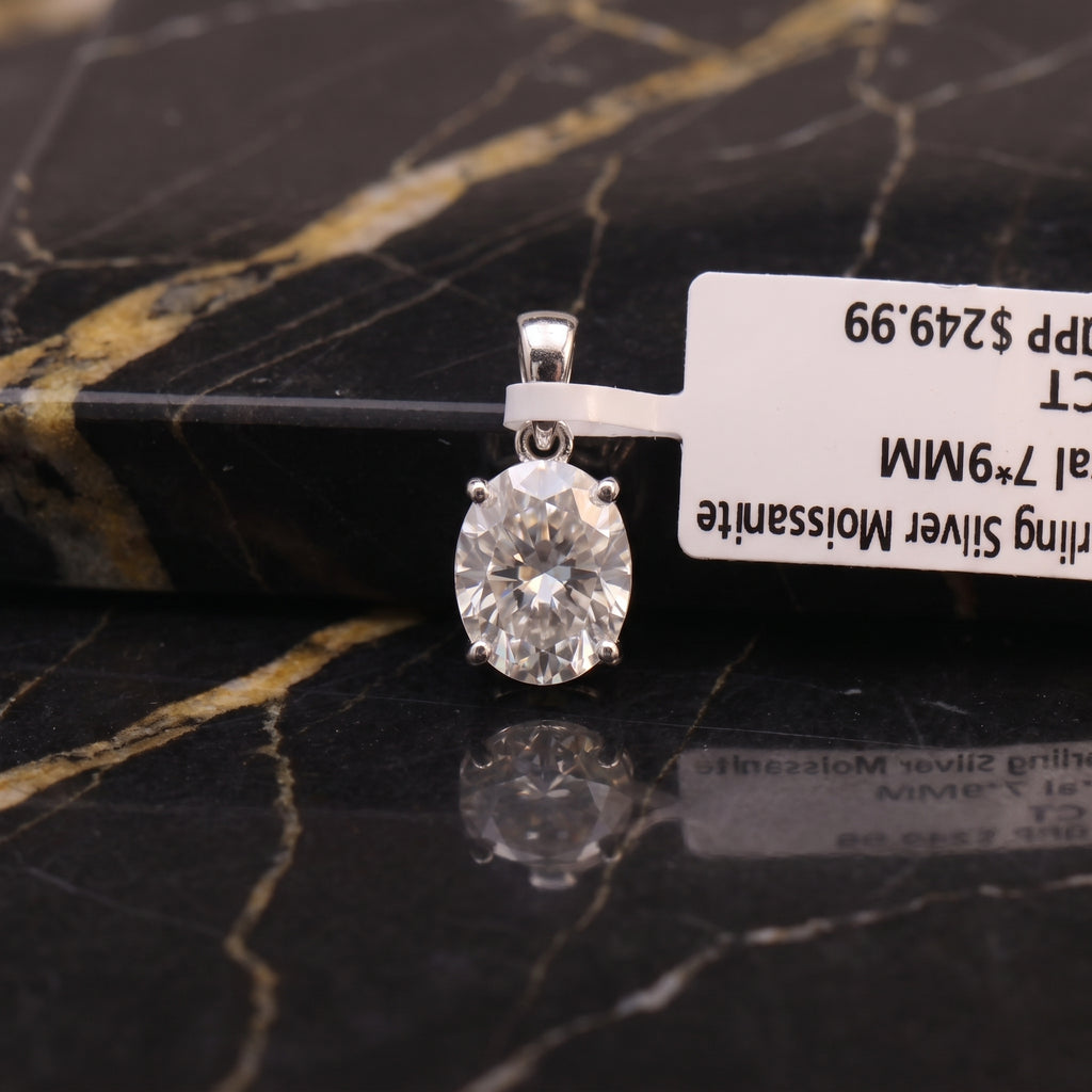 A silver pendant with a round clear gemstone in a four-prong setting, resting on a black and gold veined marble surface next to a white product tag.