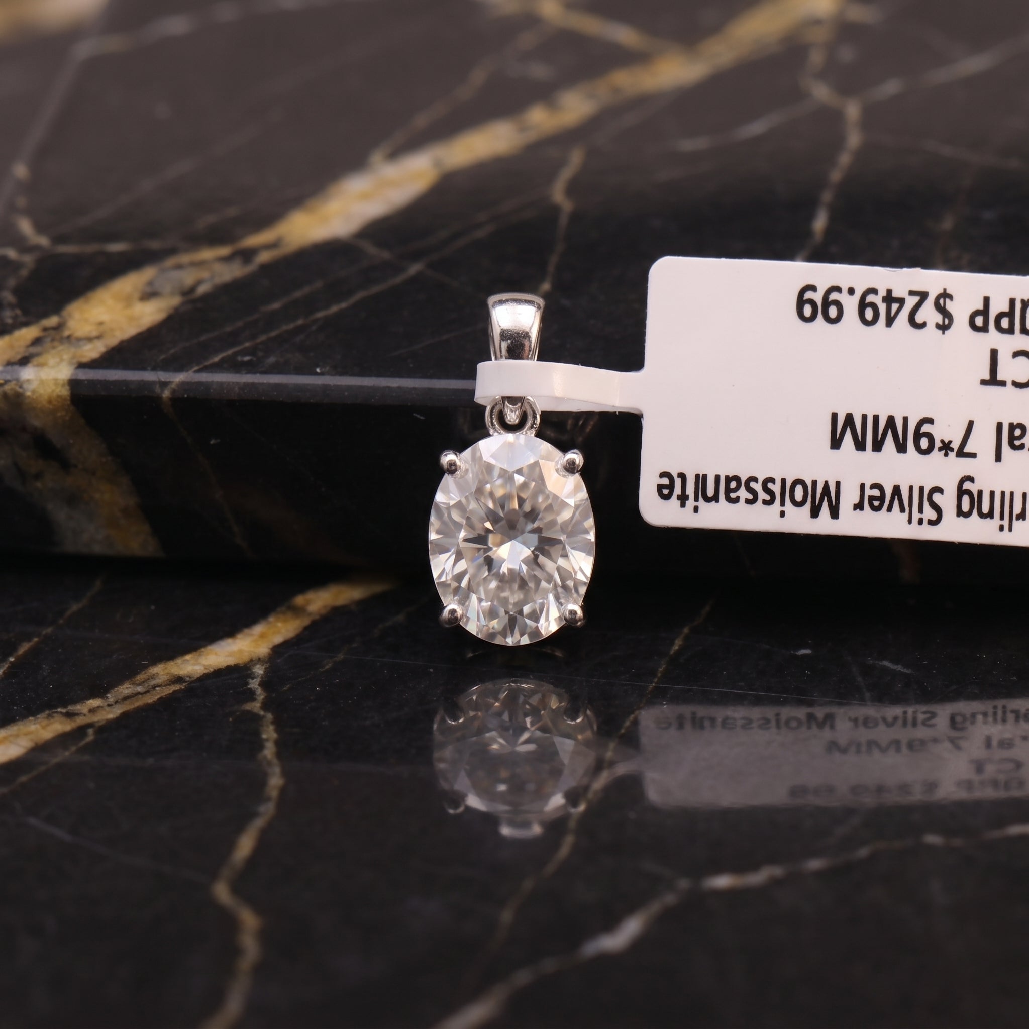 A silver pendant with a round clear gemstone in a four-prong setting, resting on a black and gold veined marble surface next to a white product tag.