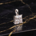 A silver pendant with a princess-cut clear gemstone, displayed on a black marble surface with gold veining.