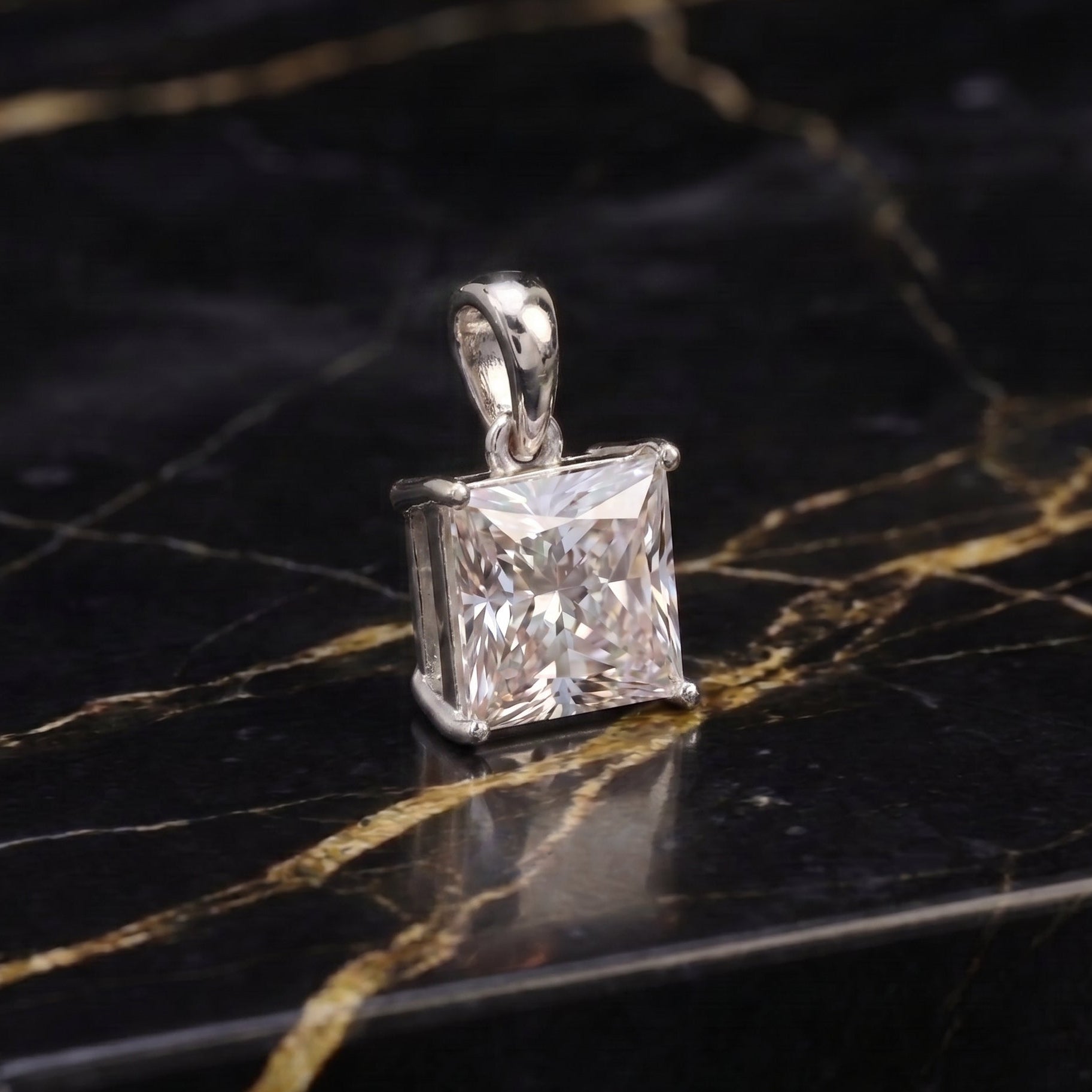A silver pendant with a princess-cut clear gemstone, displayed on a black marble surface with gold veining.