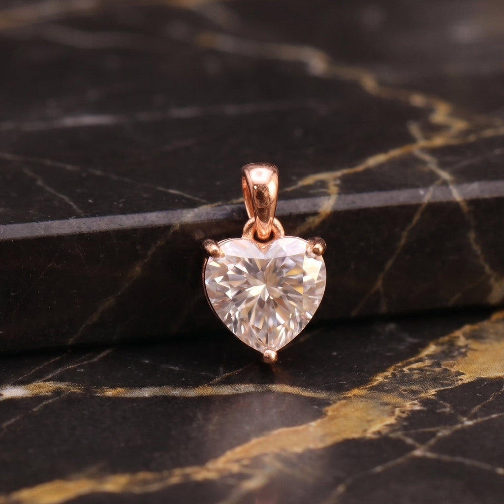 A heart-shaped clear gemstone pendant in a rose gold setting, displayed on a black and gold veined marble surface.