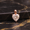 A heart-shaped clear gemstone pendant in a rose gold setting, displayed on a black and gold veined marble surface.