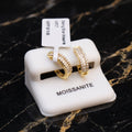 A pair of gold hoop earrings with rows of clear pavé stones, displayed on a white MOISSANITE jewelry stand over a black marble surface.