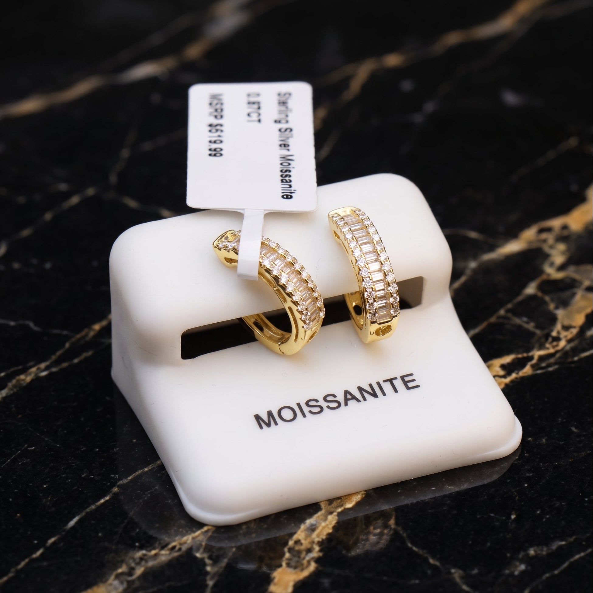 A pair of gold hoop earrings with rows of clear pavé stones, displayed on a white MOISSANITE jewelry stand over a black marble surface.
