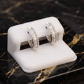A pair of silver hoop earrings covered in clear pavé stones, displayed on a white SSANITE stand set on a black and gold marble slab.