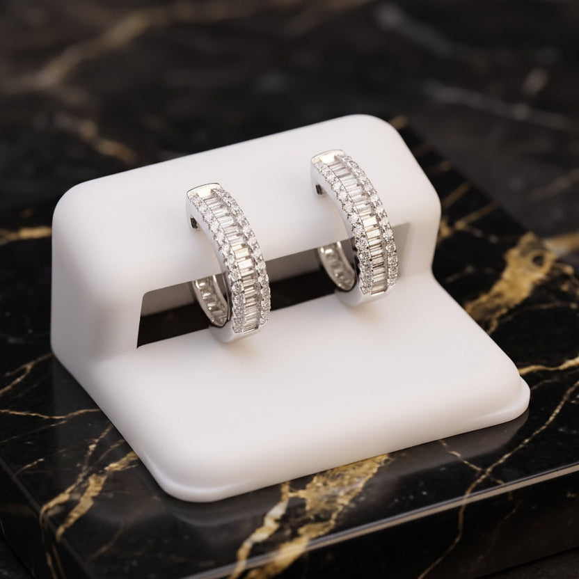 A pair of silver hoop earrings covered in clear pavé stones, displayed on a white SSANITE stand set on a black and gold marble slab.