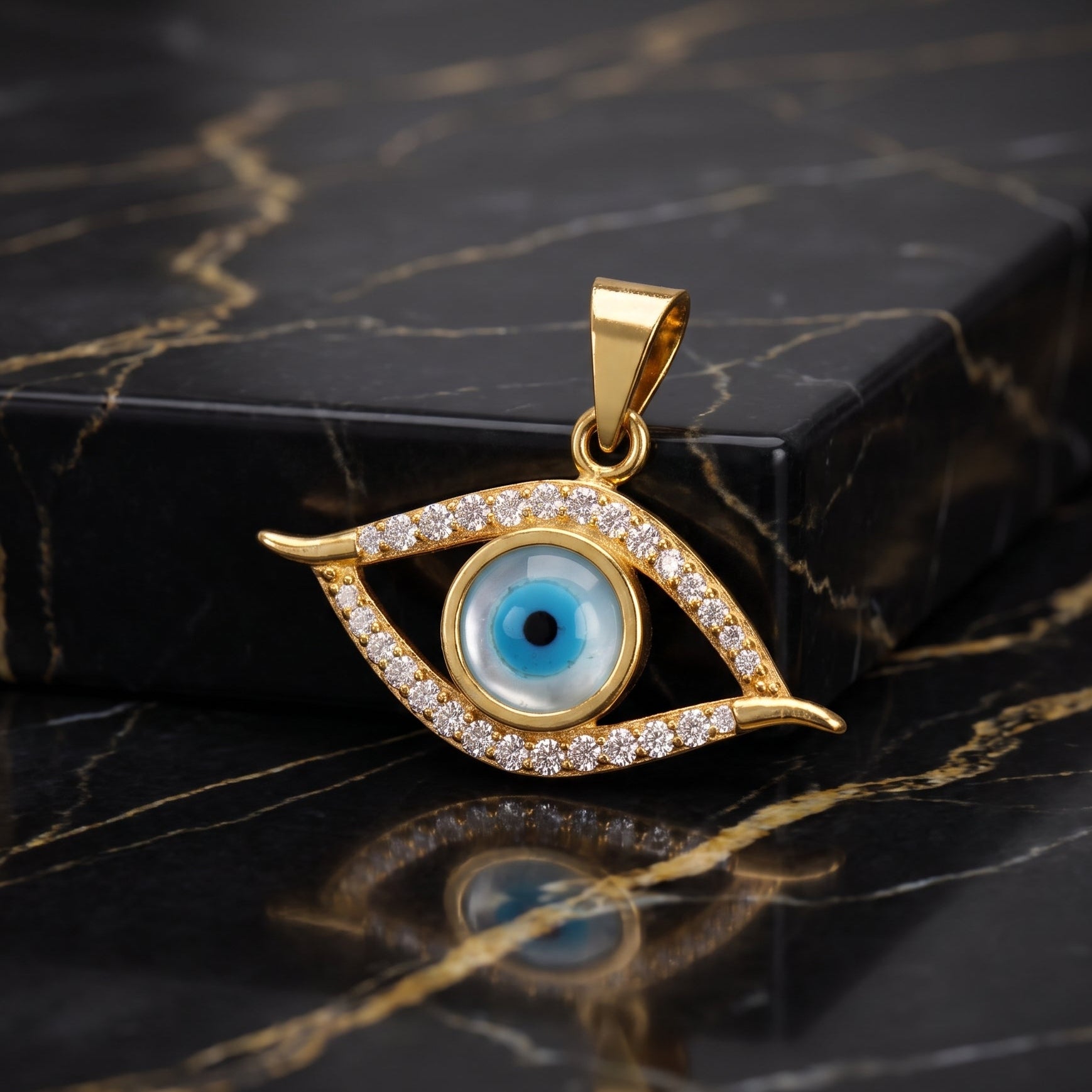 Gold evil eye pendant with a blue and black glass eye center and clear crystal accents, displayed on a black marble surface next to a black box.