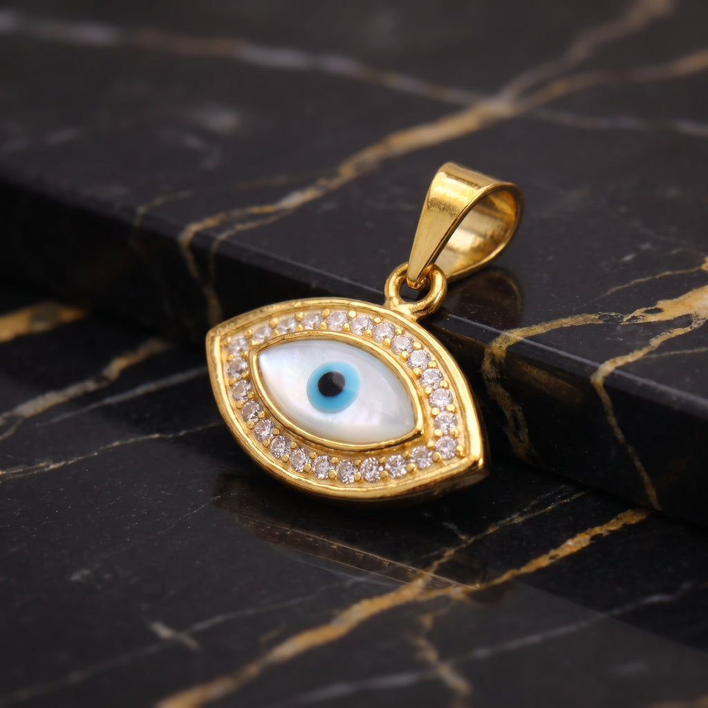 Gold evil eye pendant with a white and blue enamel center and small clear stones around the border, displayed on a black marble surface with gold veining.