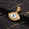 Gold evil eye pendant with a white and blue enamel center and small clear stones around the border, displayed on a black marble surface with gold veining.