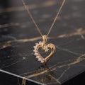 A gold heart-shaped pendant necklace with a row of small clear stones along one side, displayed on a black marble surface.